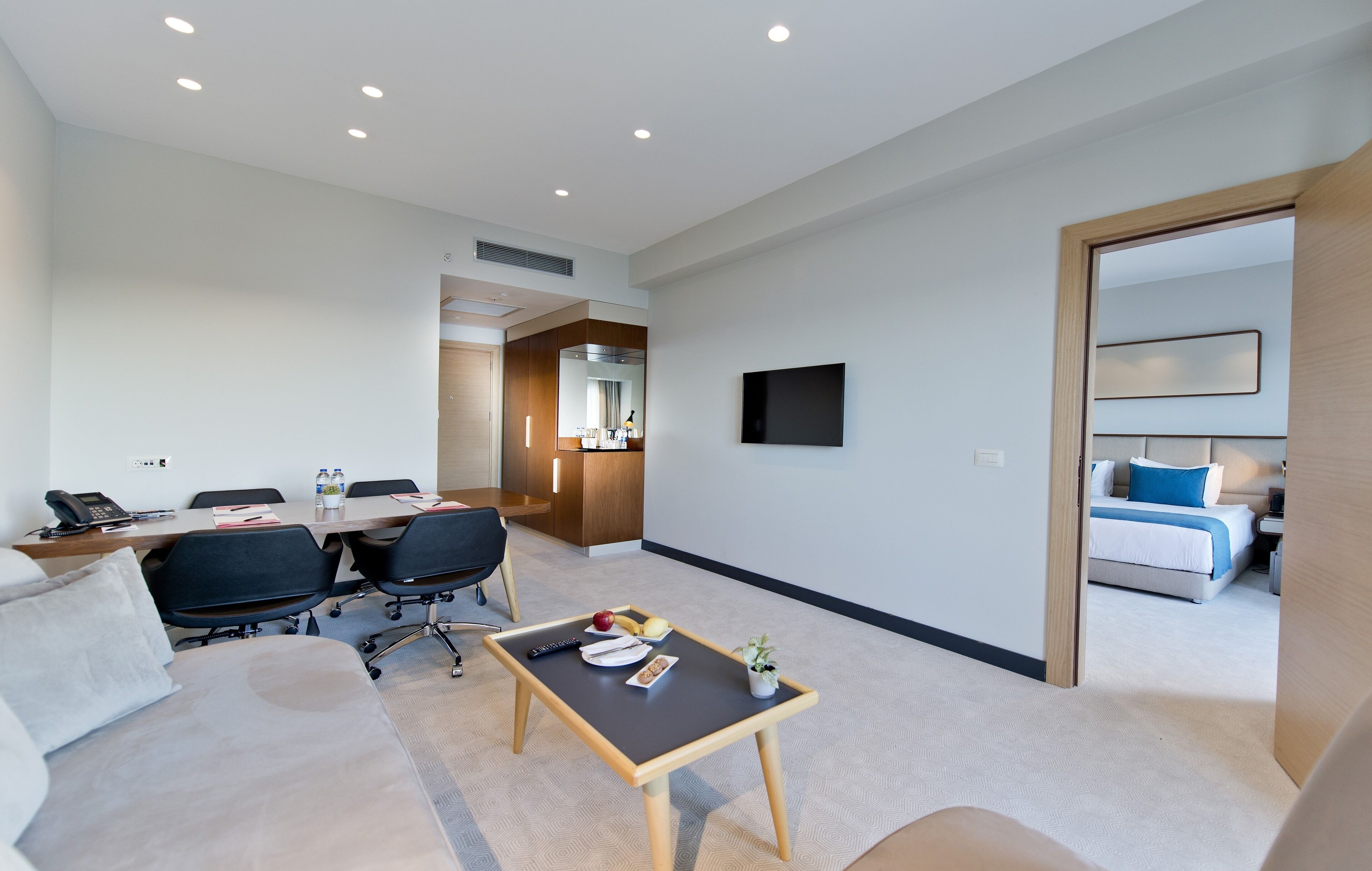 A modern hotel room with a large bed, a small desk with a chair, a sofa, a TV mounted on the wall, and a dining area with a table and chairs.