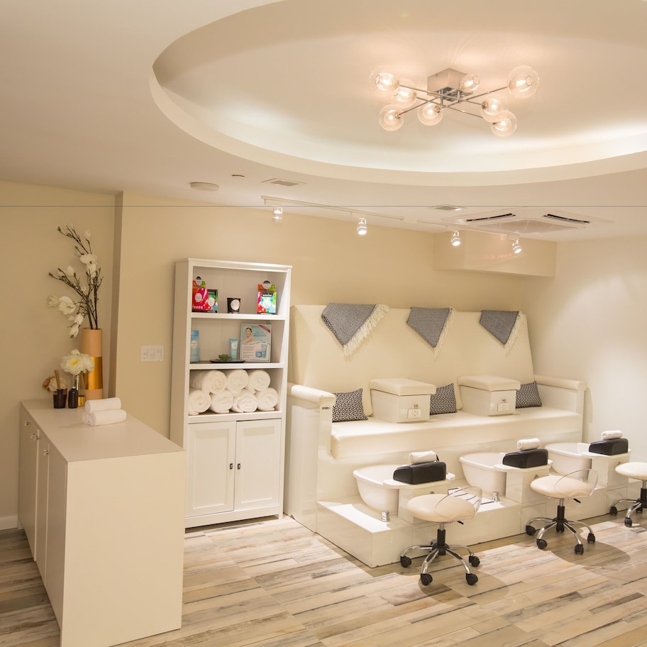 A modern spa room with a reception desk, a shelf with toiletries, a seating area with massage chairs, and a ceiling light fixture.