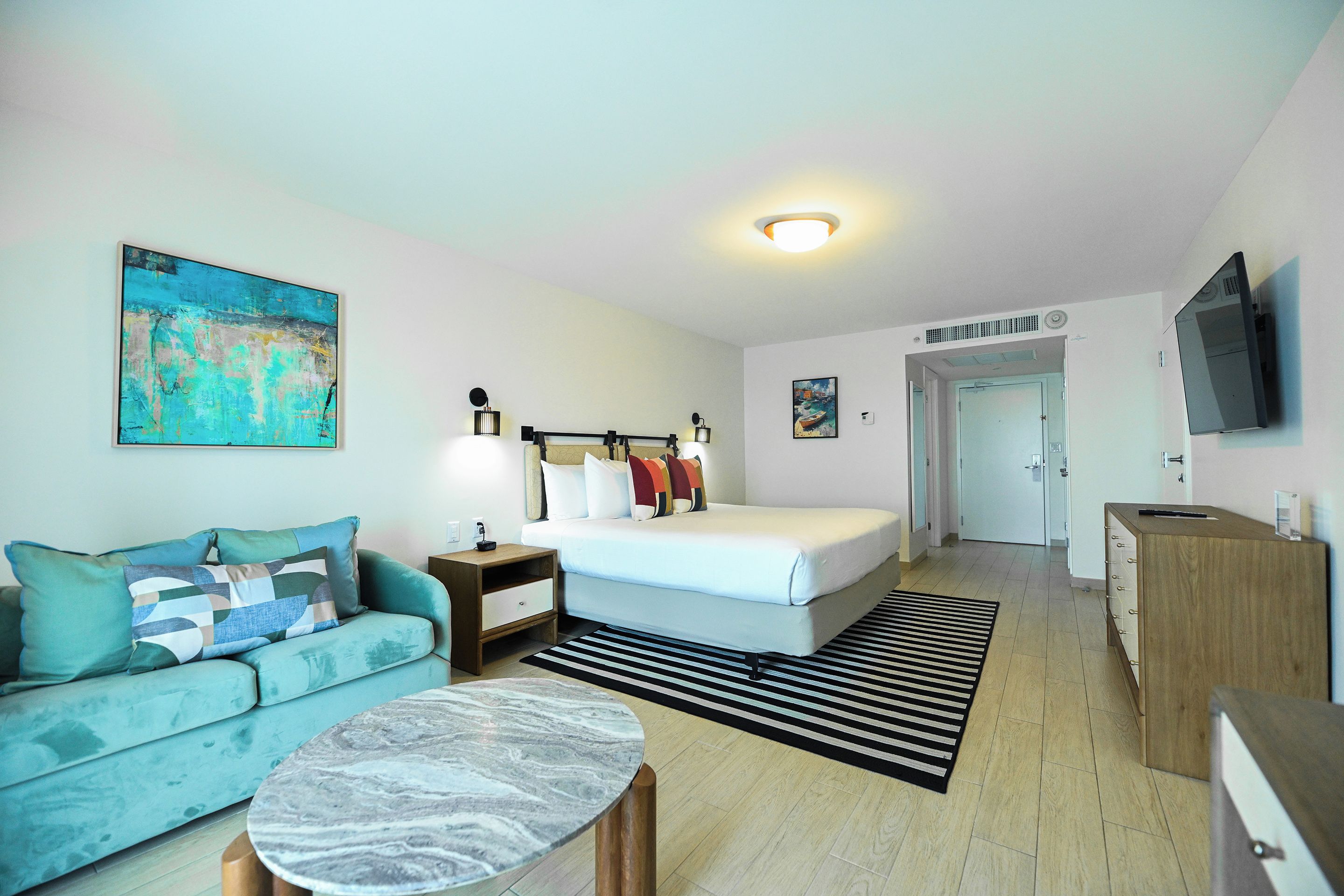 Una habitación de hotel moderna con una cama, un sofá, una mesita de café, un cuadro y un televisor.