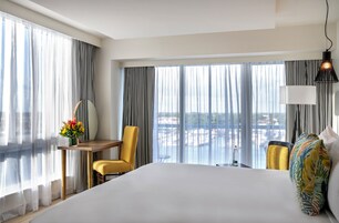 Una habitación de hotel con una cama grande, un escritorio, dos sillas, vistas de la ciudad a través de amplias ventanas y un balcón.
