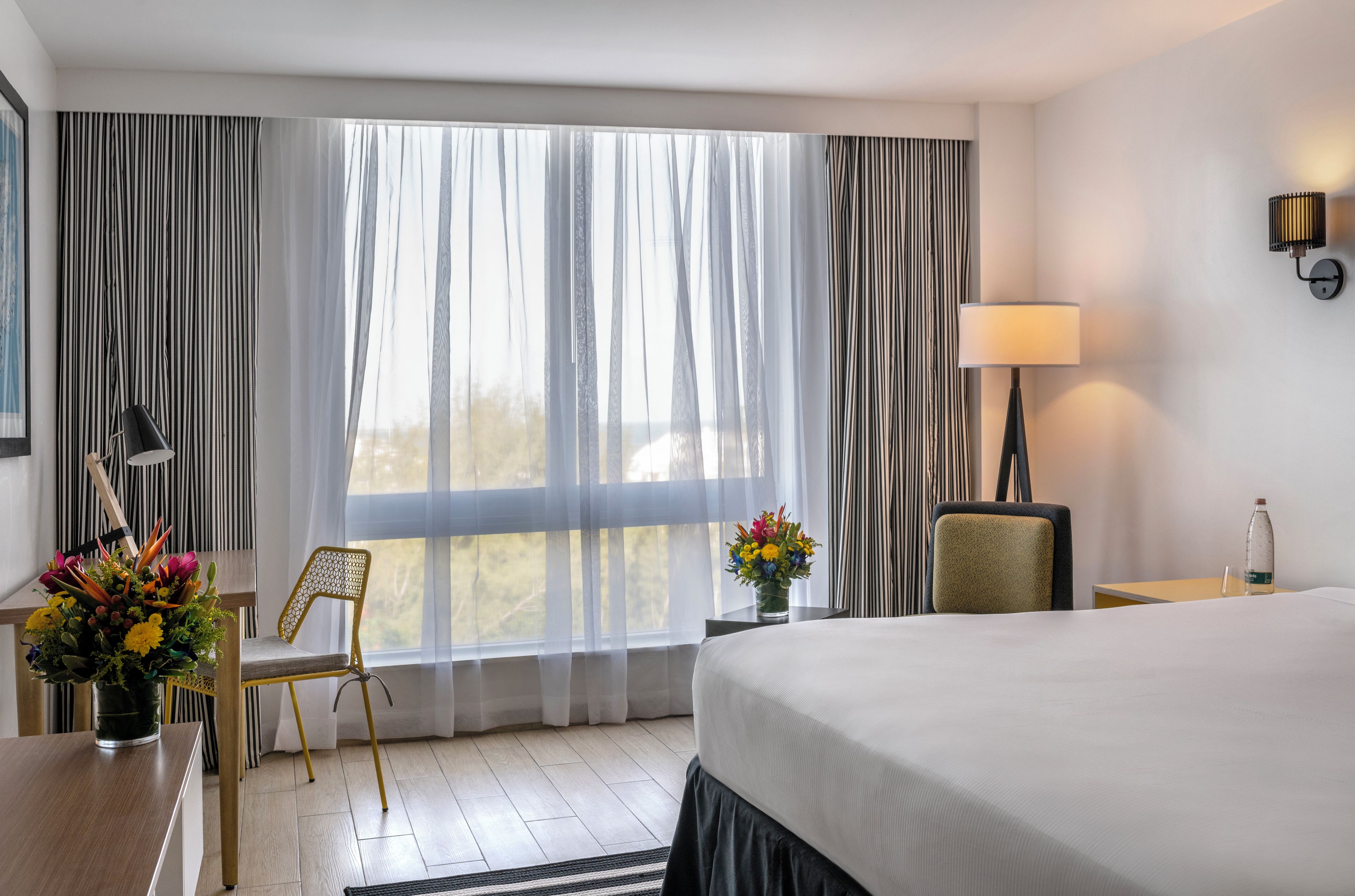 Una habitación de hotel con cama, escritorio con lámpara, silla, jarrón con flores y ventana con cortinas.