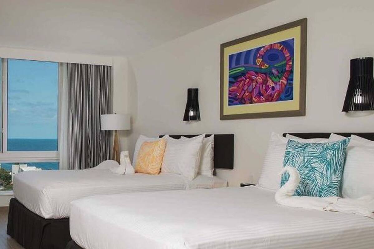 Habitación de hotel con dos camas, un ventanal con cortinas, un cuadro en la pared y una lámpara a cada lado de las camas.