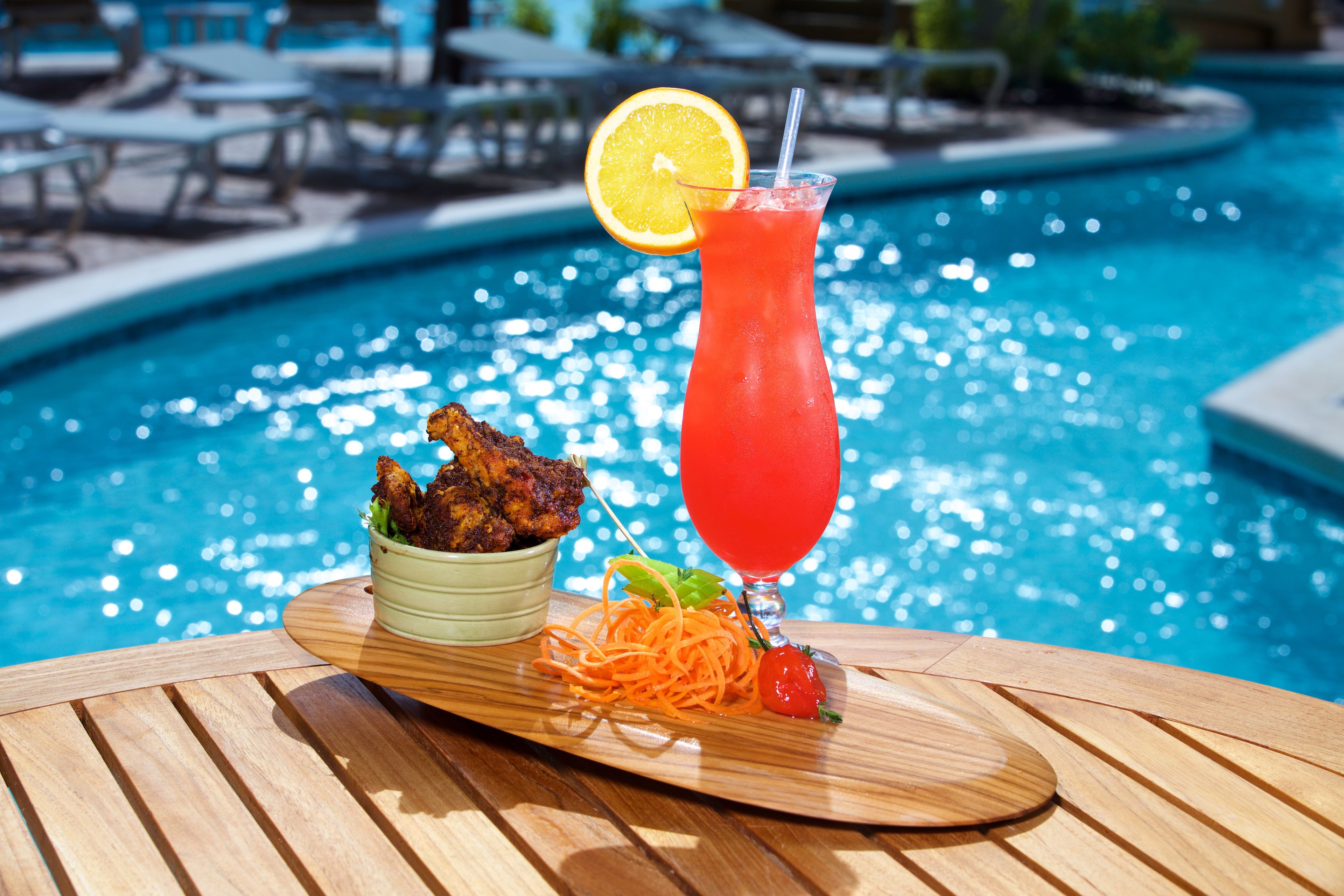 Une table à manger dressée au bord de la piscine, avec un repas composé de poulet frit, d’une salade et d’une boisson rouge garnie d’une tranche de citron.