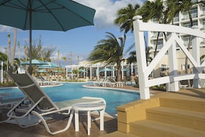 A pool area with lounge chairs and umbrellas, surrounded by palm trees and a building with balconies. - Warwick Paradise Island- All Inclusive- Adults Only (Paradise Island)