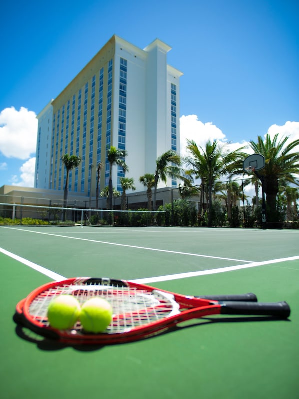 A tennis court with a red and black tennis racket and three tennis balls in the foreground, a net, and palm trees in the background.