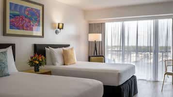 A hotel room with two beds, a nightstand, a lamp, a painting, and a view of the city through large windows.