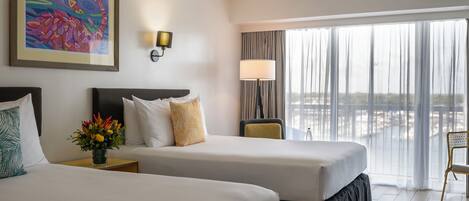 A hotel room with two beds, a nightstand, a lamp, a painting, and a view of the city through large windows.