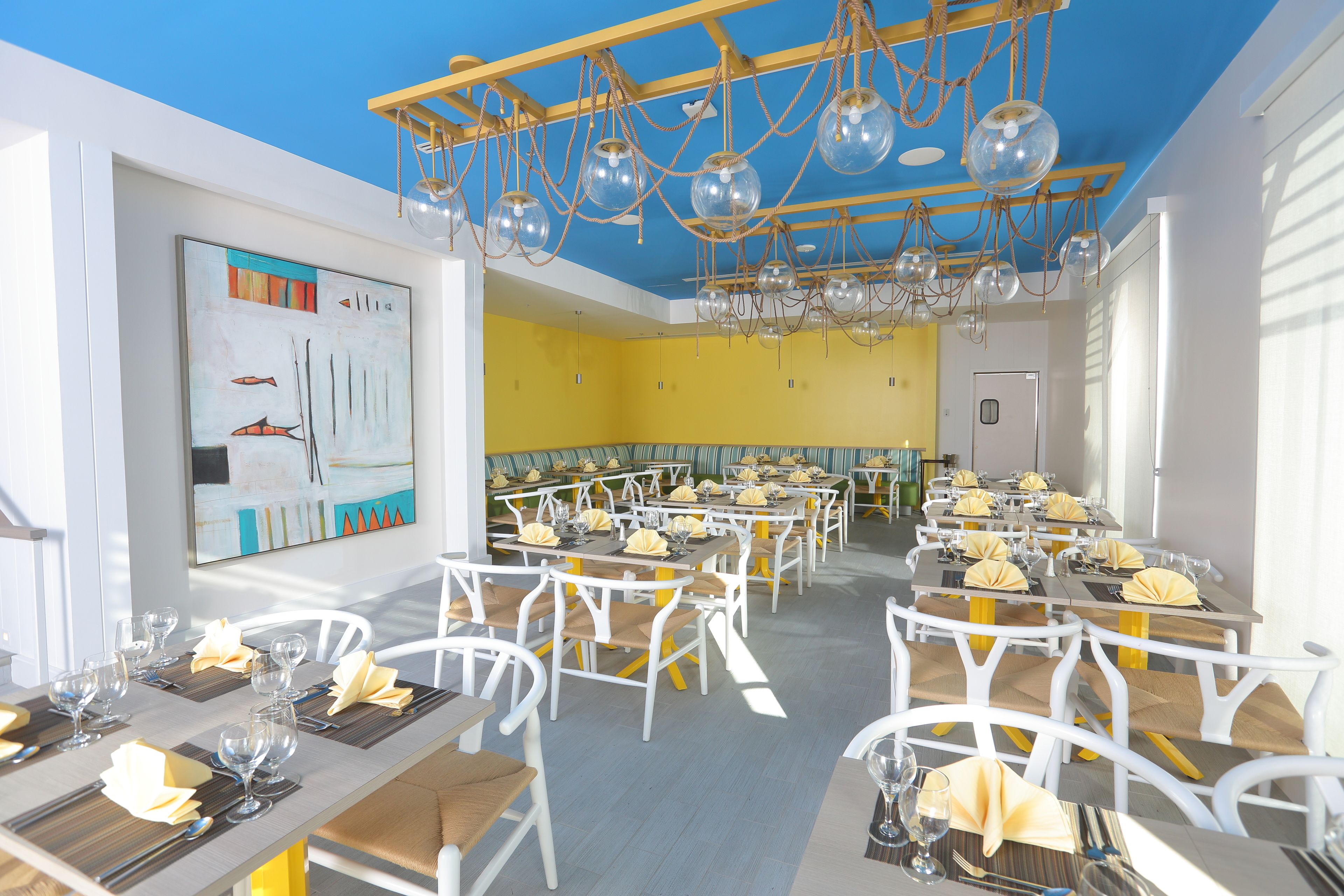 Un ristorante con sedie e tavoli bianchi, tovaglioli gialli e un soffitto blu decorato con globi di vetro sospesi.