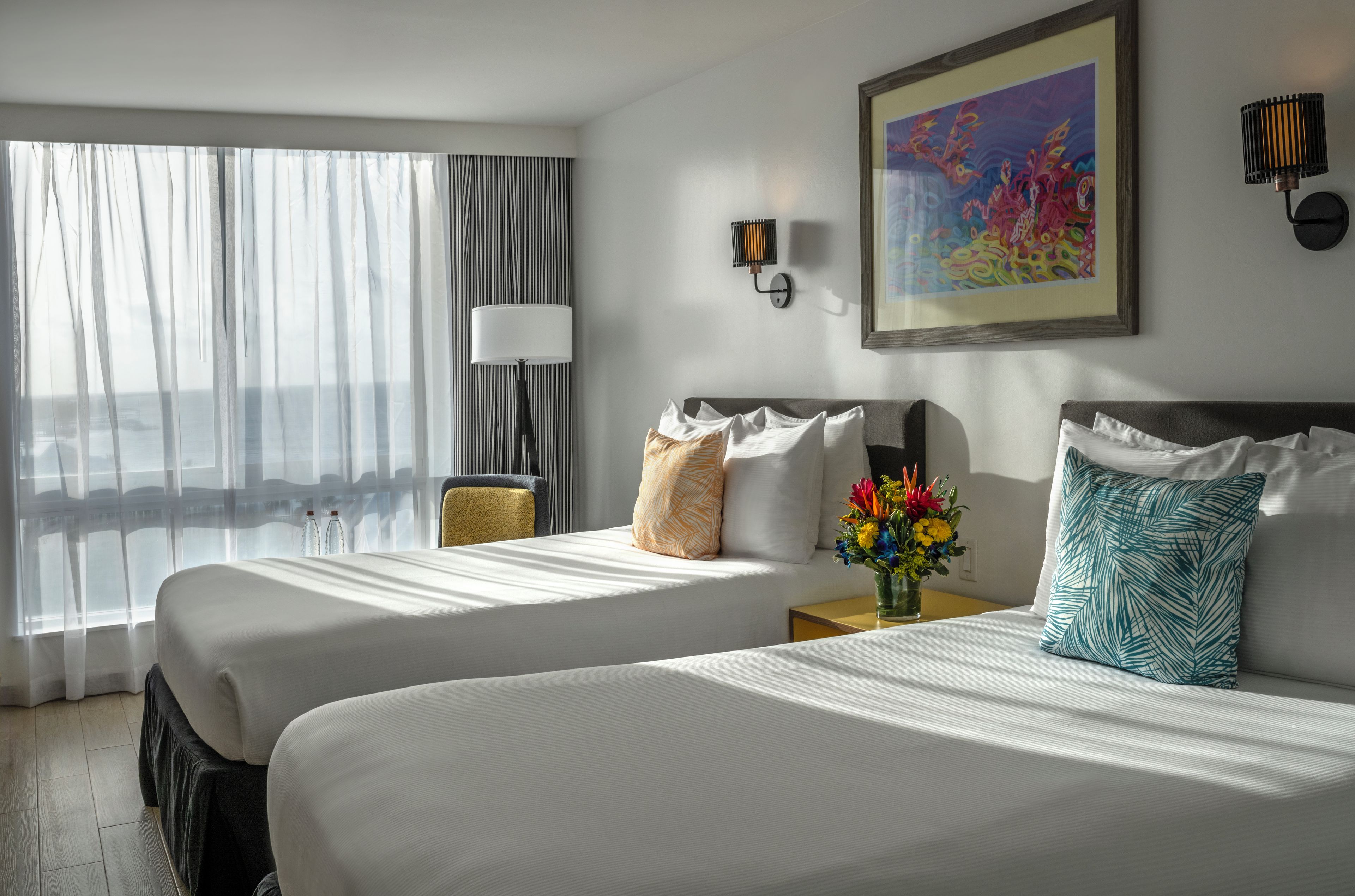 Une chambre d’hôtel avec deux lits, une grande fenêtre avec des rideaux, un tableau au mur et une table de chevet avec une lampe et un vase de fleurs.