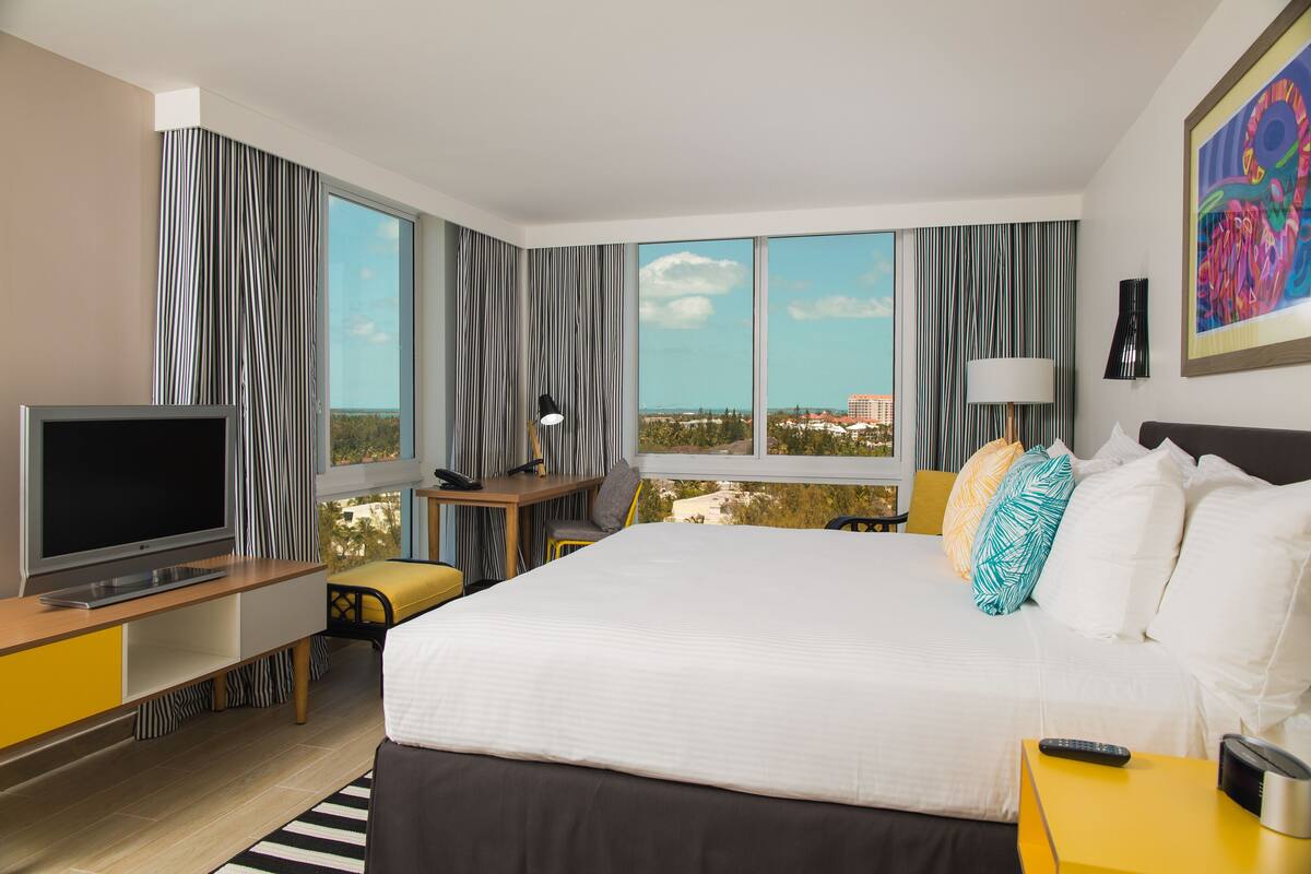 Habitación de hotel con una cama grande, un escritorio con televisor, una silla, una lámpara y vistas amplias de la ciudad.