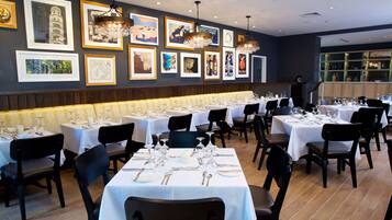 Un restaurante con varias mesas dispuestas para comer, obras de arte en las paredes y una zona de bar visible a través del cristal.