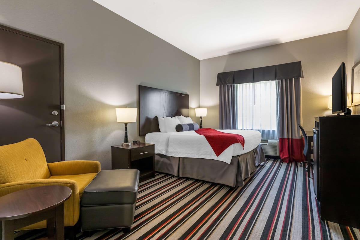 A hotel room with a bed, a yellow armchair, a small table, a nightstand, a TV, and a window with curtains.