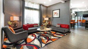 A modern hotel room with a grey sofa, armchair, and a small coffee table. There is a dining area with a bar and chairs.