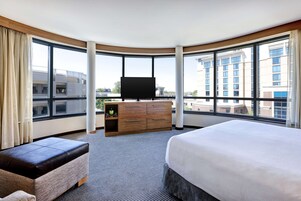 Habitación de hotel con una cama grande, un aparador de madera, un televisor de pantalla plana y vistas de la ciudad a través de amplias ventanas.