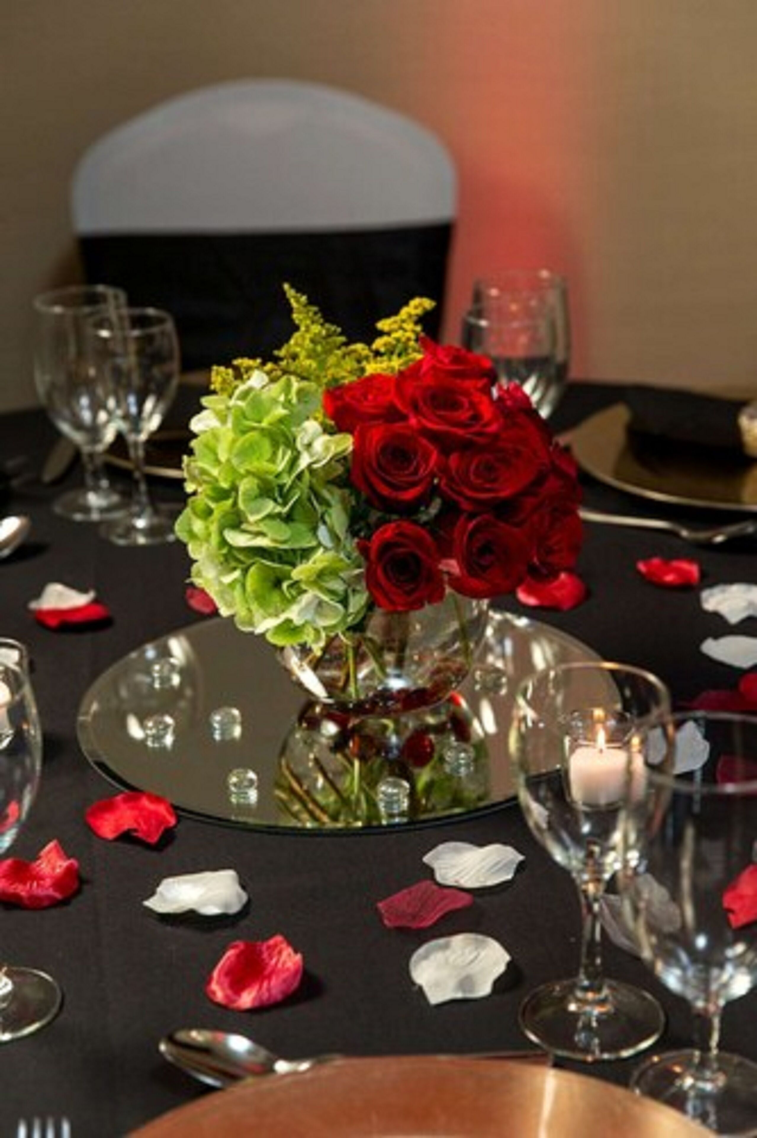 A table set for a formal event with a centerpiece of red roses and green foliage, surrounded by wine glasses and a candle.