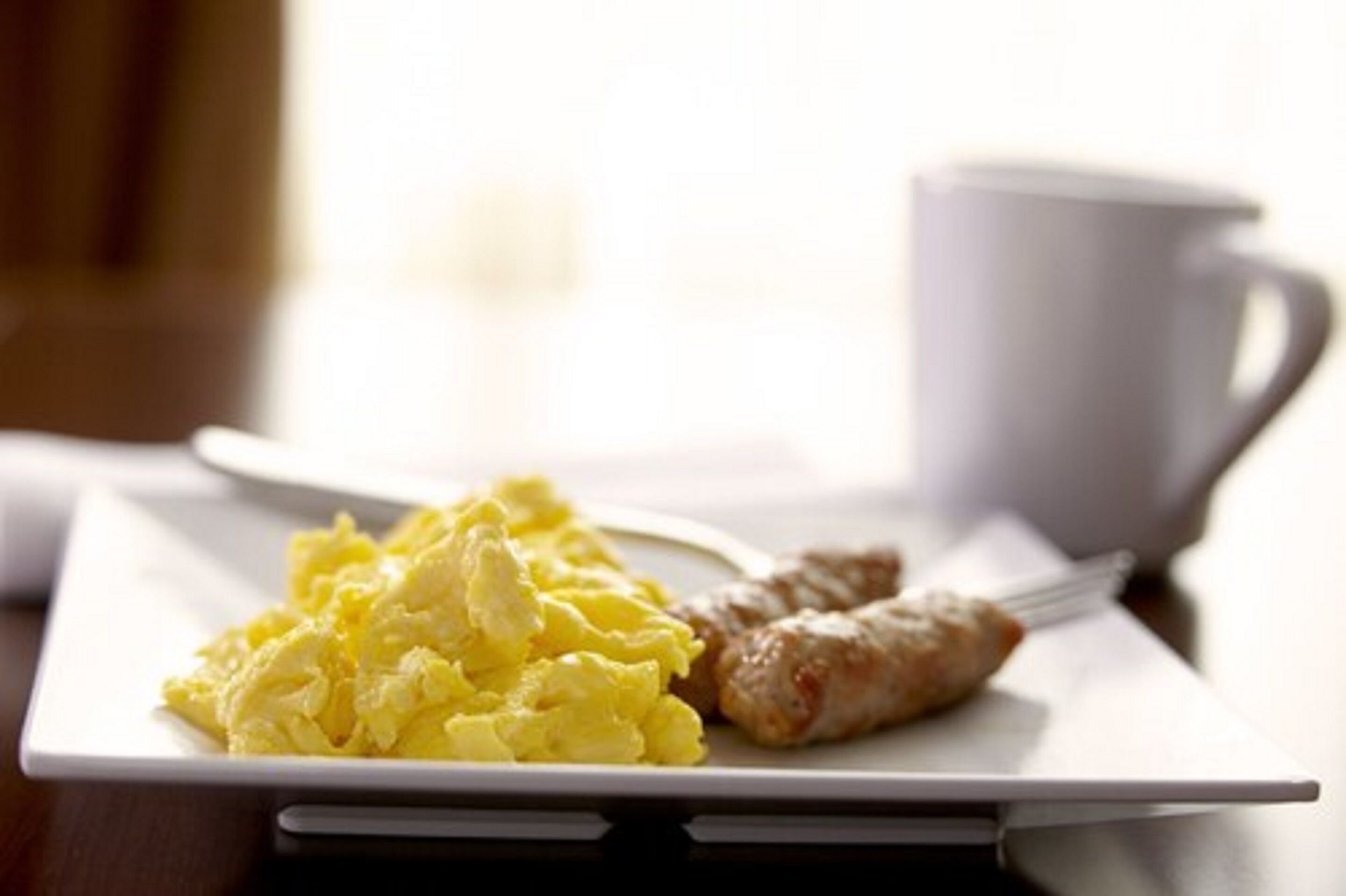 Une assiette de petit-déjeuner avec des œufs brouillés et des saucisses, une tasse blanche et une tablette posée sur une table.