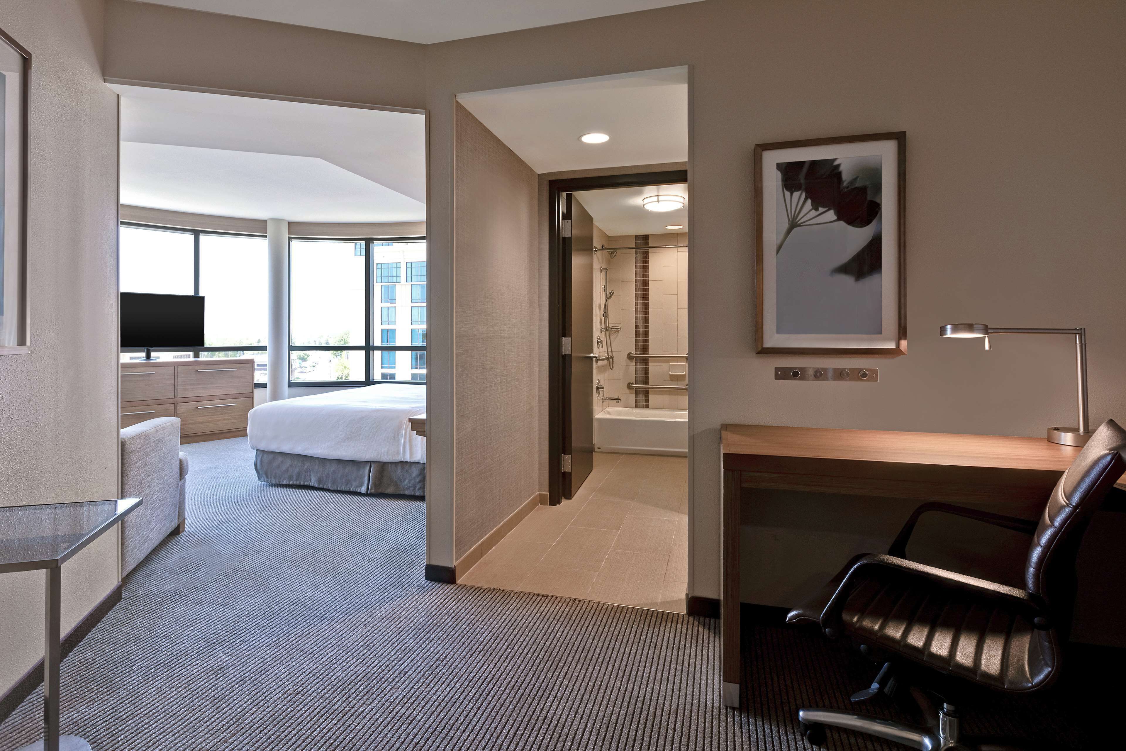 Une chambre d’hôtel équipée d’un lit, d’un bureau, d’une chaise, d’une télévision et d’une salle de bain avec douche, ainsi que d’une grande fenêtre donnant sur la ville.