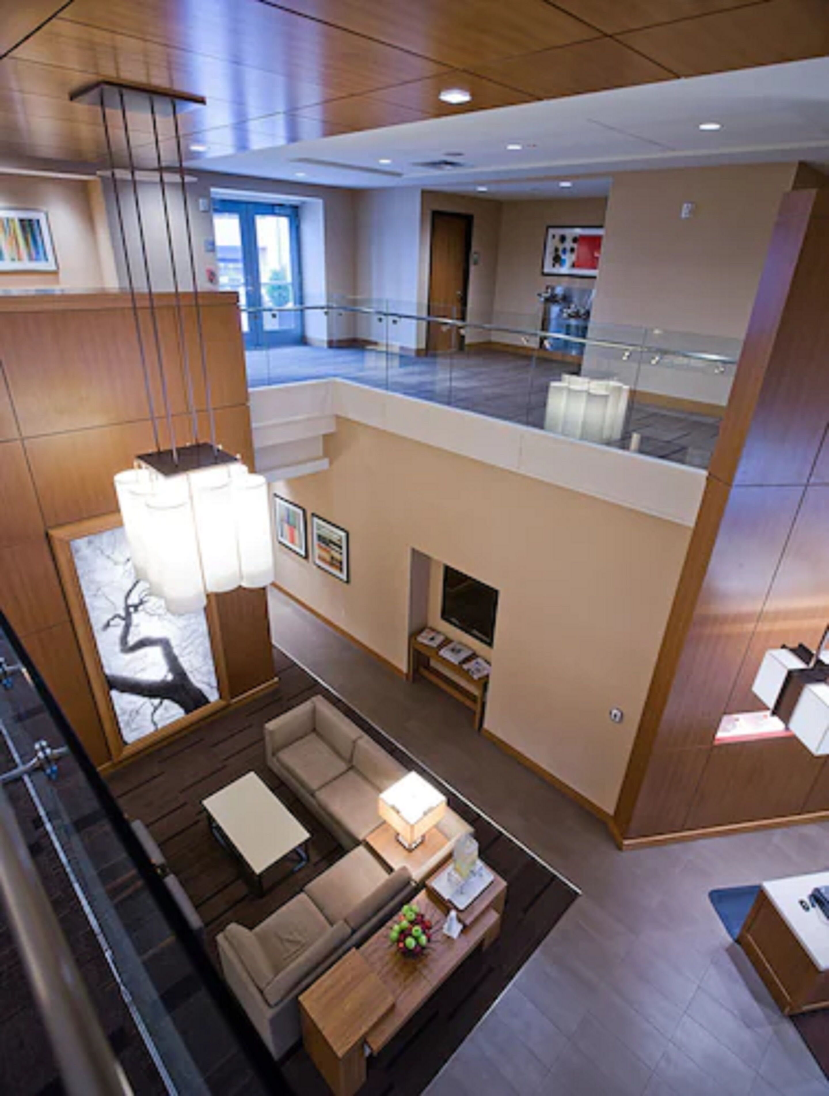 A spacious hotel lobby with a high ceiling, a central seating area with a sofa and coffee table, a fireplace, and a view of the upper floor through a glass railing.