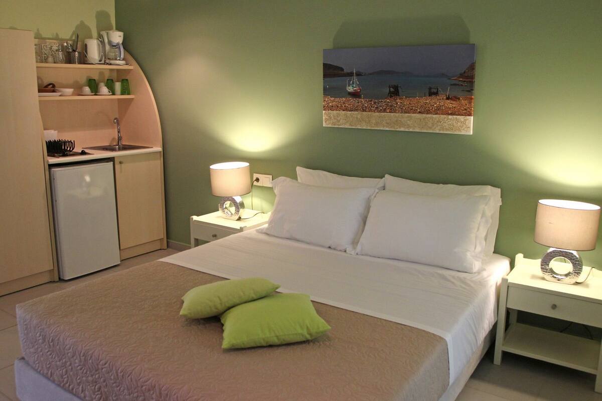 A hotel room with a bed, two bedside tables with lamps, a small kitchenette, and a painting on the wall.