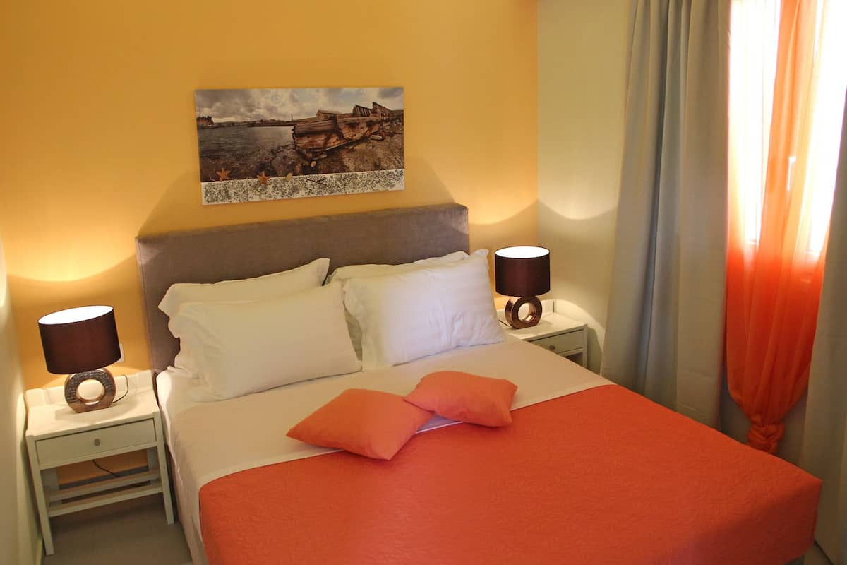 A hotel room with a bed, two bedside tables, a nightstand, a lamp, a painting, and a window with curtains.