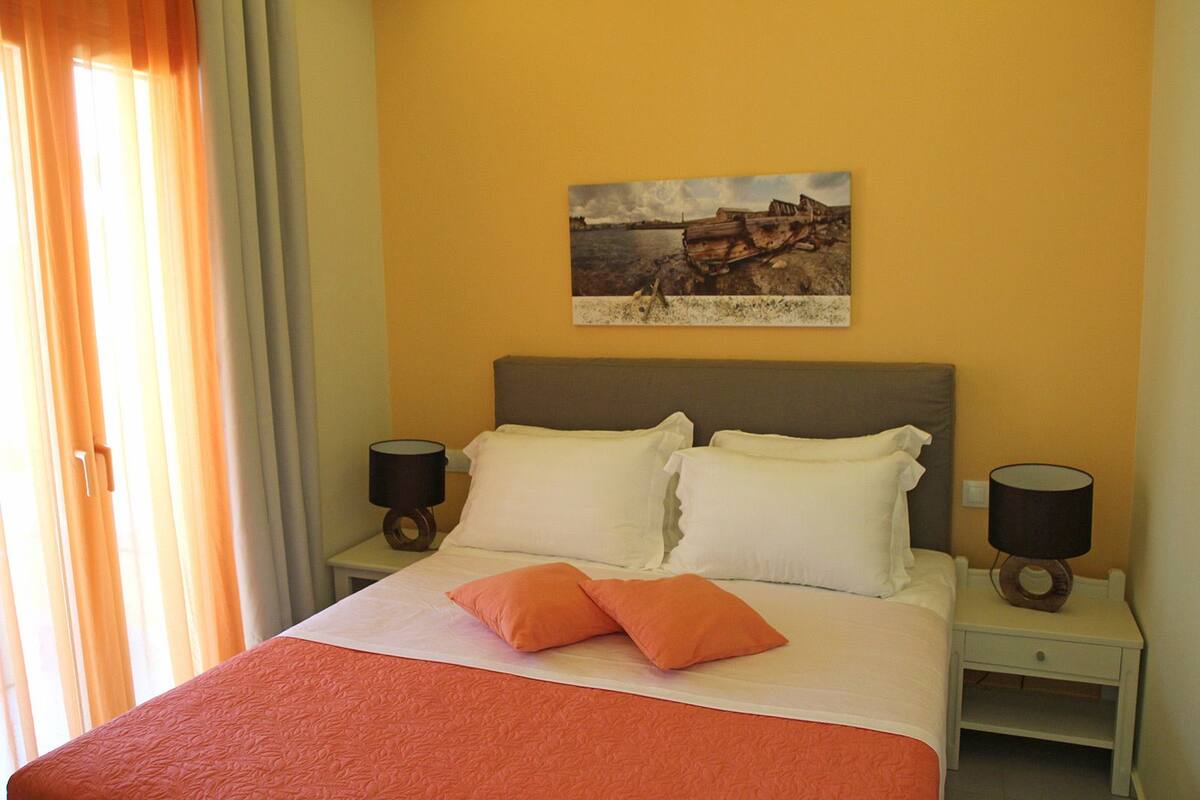 A hotel room with a bed, two bedside tables, a nightstand, a painting on the wall, and a window with curtains.