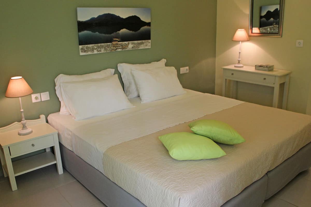 A neatly made bed with white linens and two green pillows, a bedside lamp, and a painting on the wall.