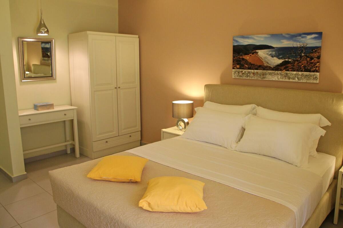 A hotel room with a bed, two pillows, a nightstand with a lamp, a wardrobe, a desk with a mirror, and a painting on the wall.