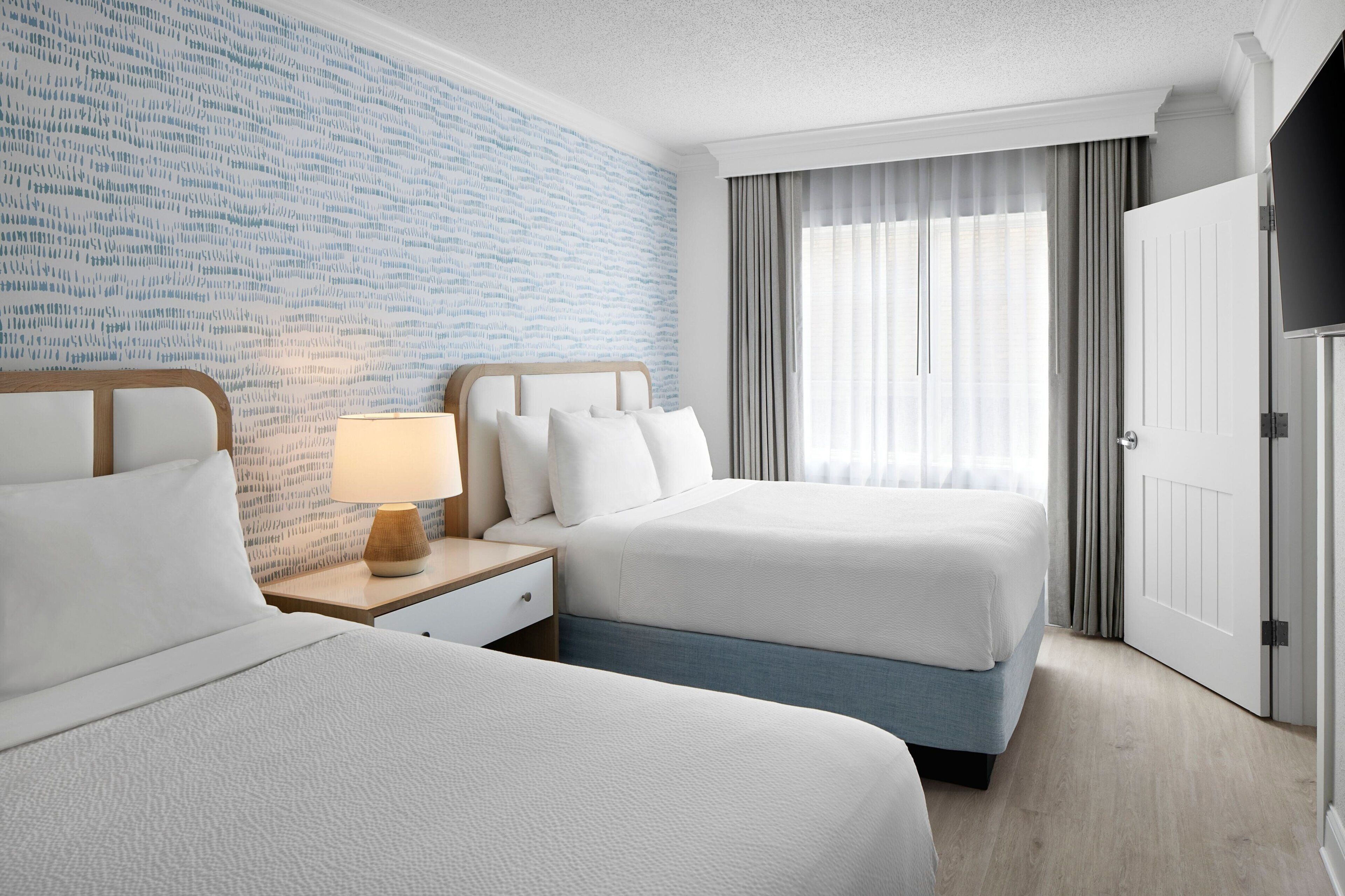 Een hotelkamer met twee bedden, een nachtkastje met lamp, een groot raam met gordijnen en een deur.