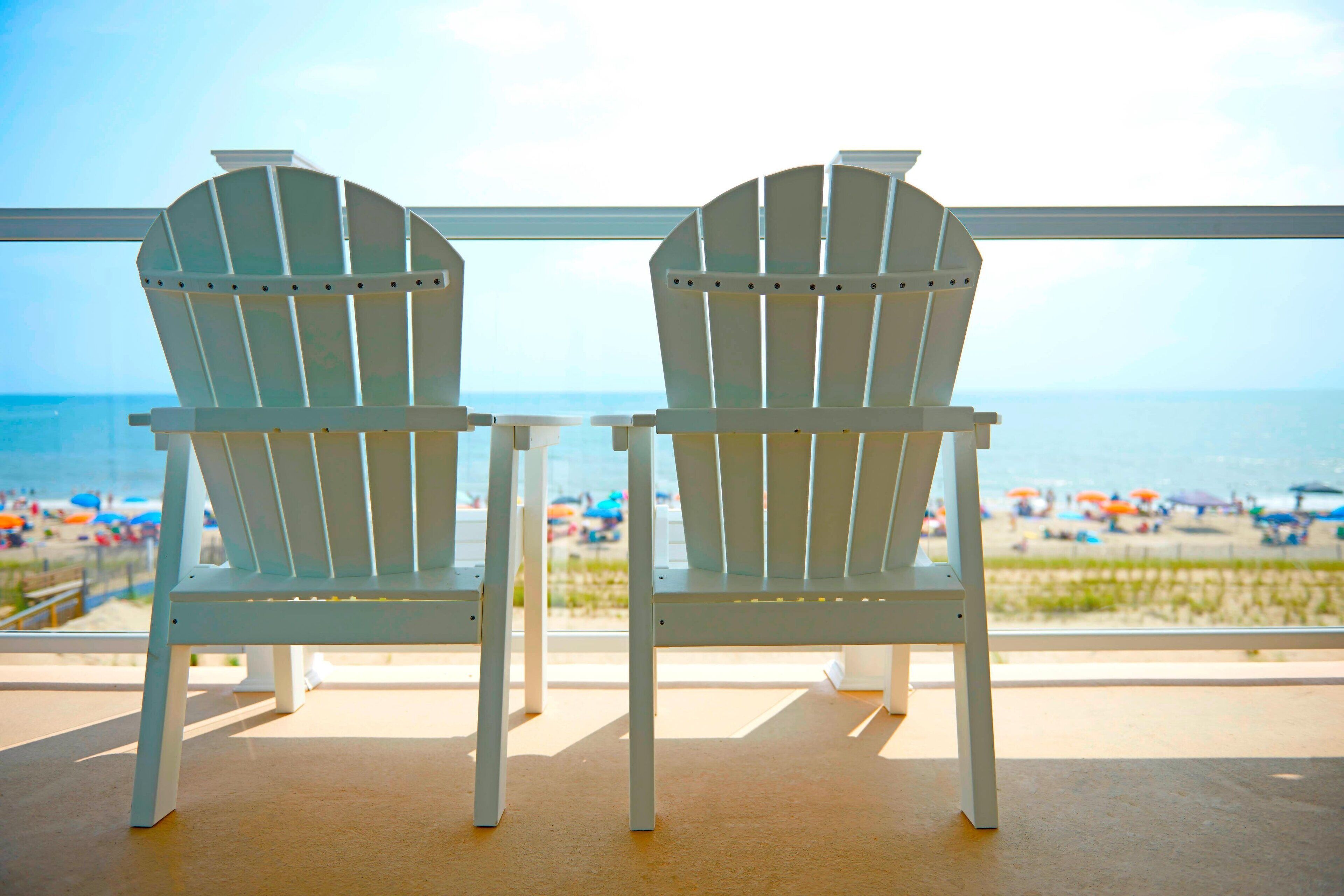 Two white Adirondack chairs facing a beach with umbrellas and people in the background.