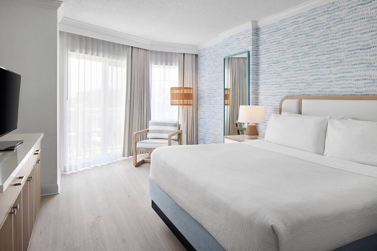 Ein Hotelzimmer mit einem großen Bett, einem Fernseher, einem Sessel, einem Nachttisch und einem Fenster mit Vorhängen.