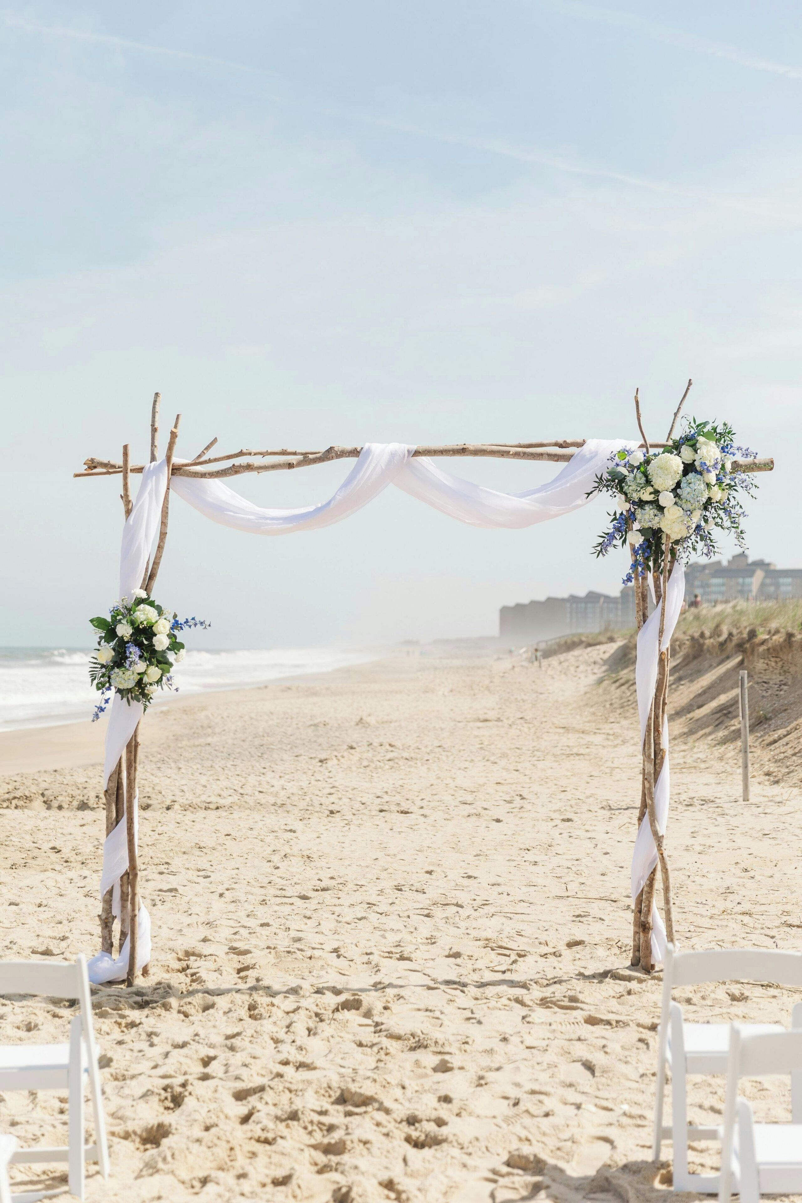 Een bruiloftstrouwboog op het strand, versierd met witte draperingen en bloemdecoraties.
