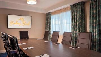 Uma sala de conferências com uma mesa comprida, seis cadeiras, uma tela de projetor exibindo "Bethany Beach Ocean Suites" e amplas janelas com cortinas estampadas.