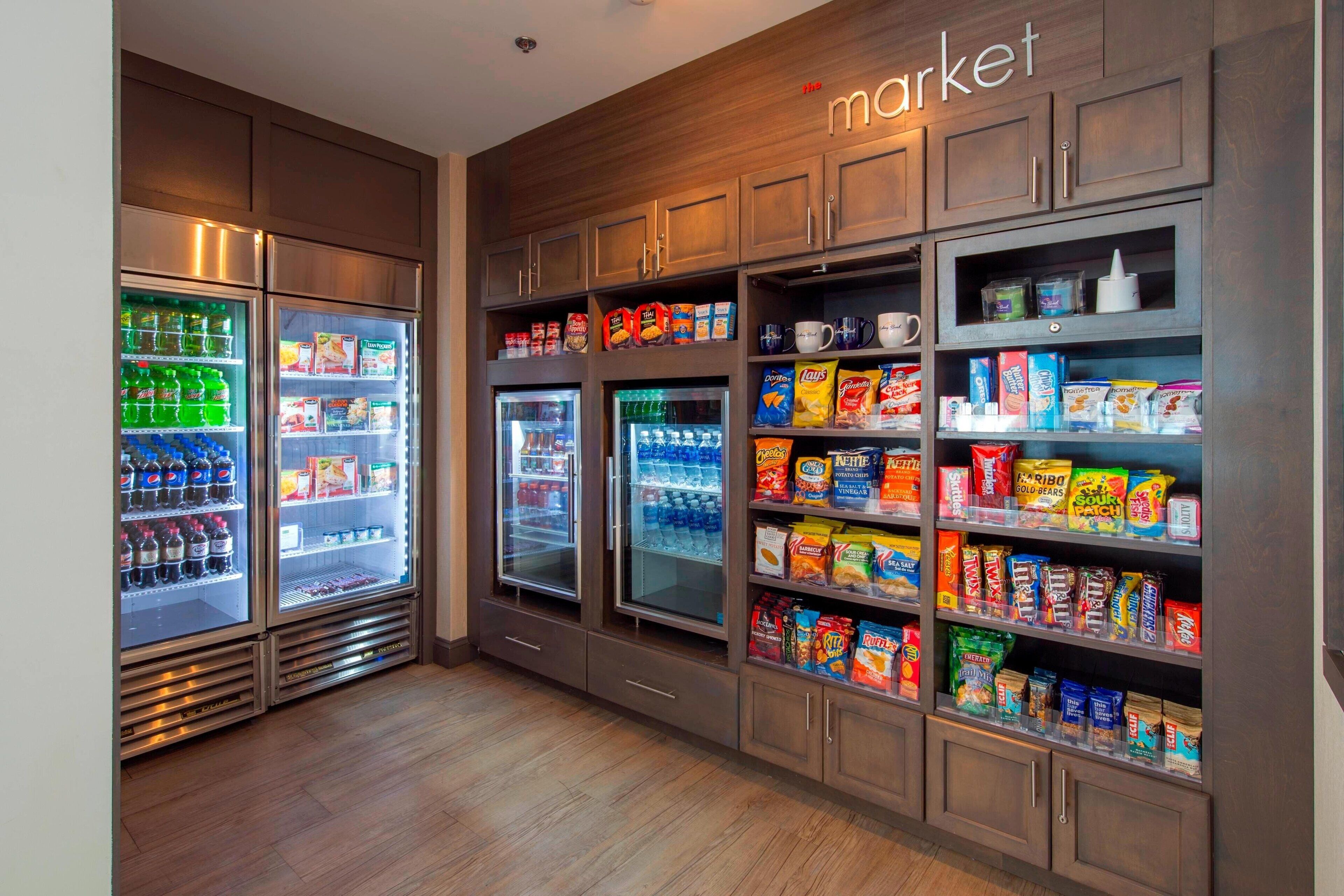 A self-service snack and beverage area with refrigerated drink dispensers and shelves stocked with various snacks and cereals.