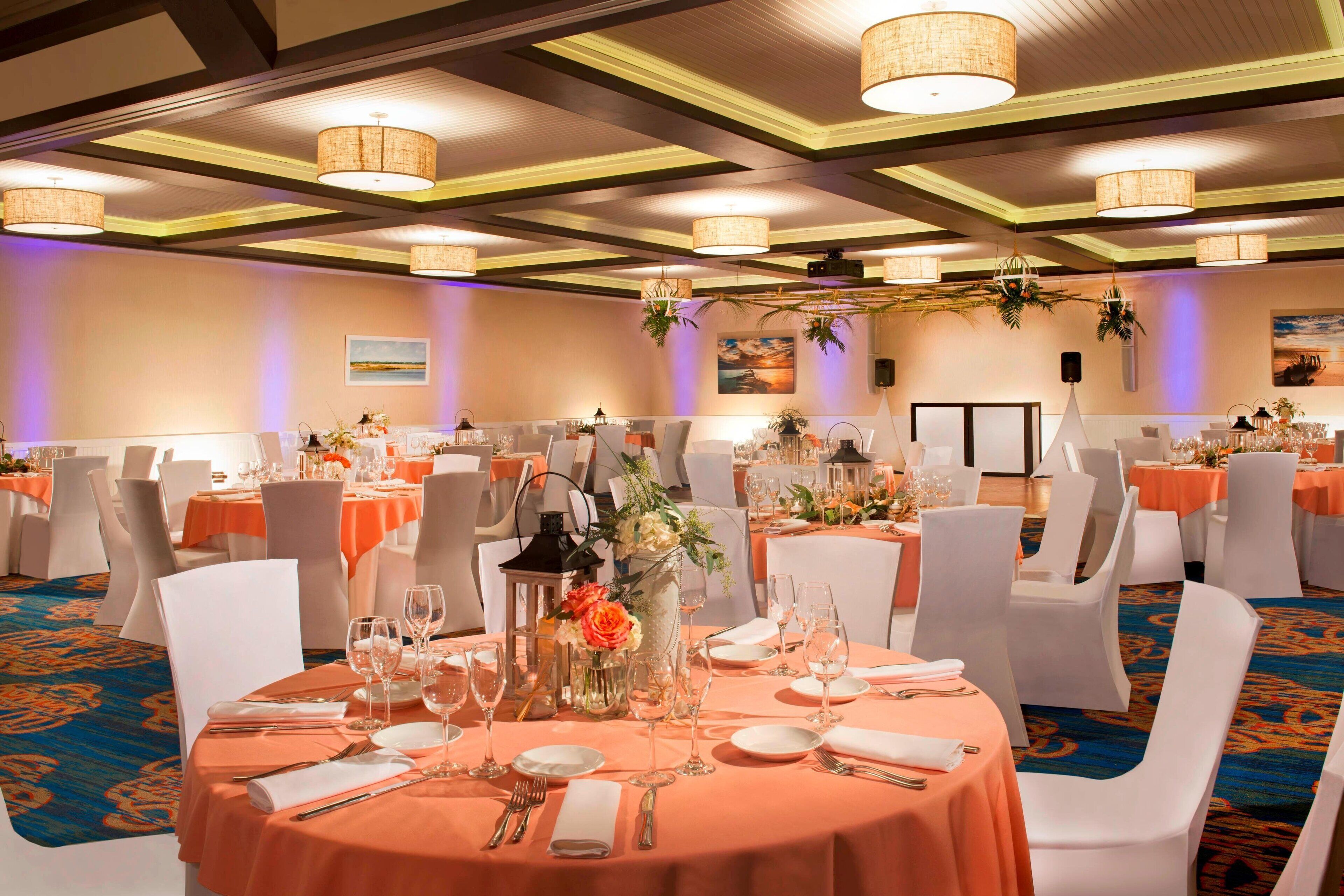 A banquet hall with round tables set for a formal event, featuring white chairs, orange tablecloths, and floral centerpieces.