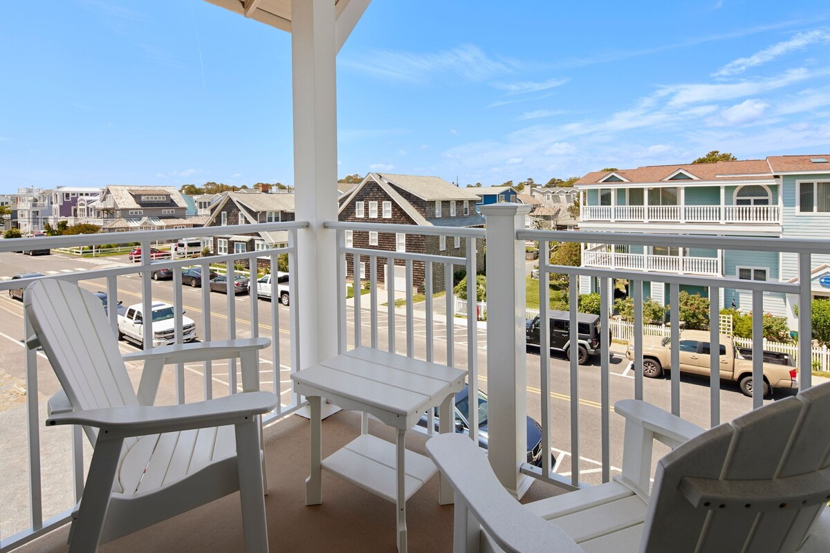 A balcony with white Adirondack chairs and a small table, overlooking a street with parked cars and houses.