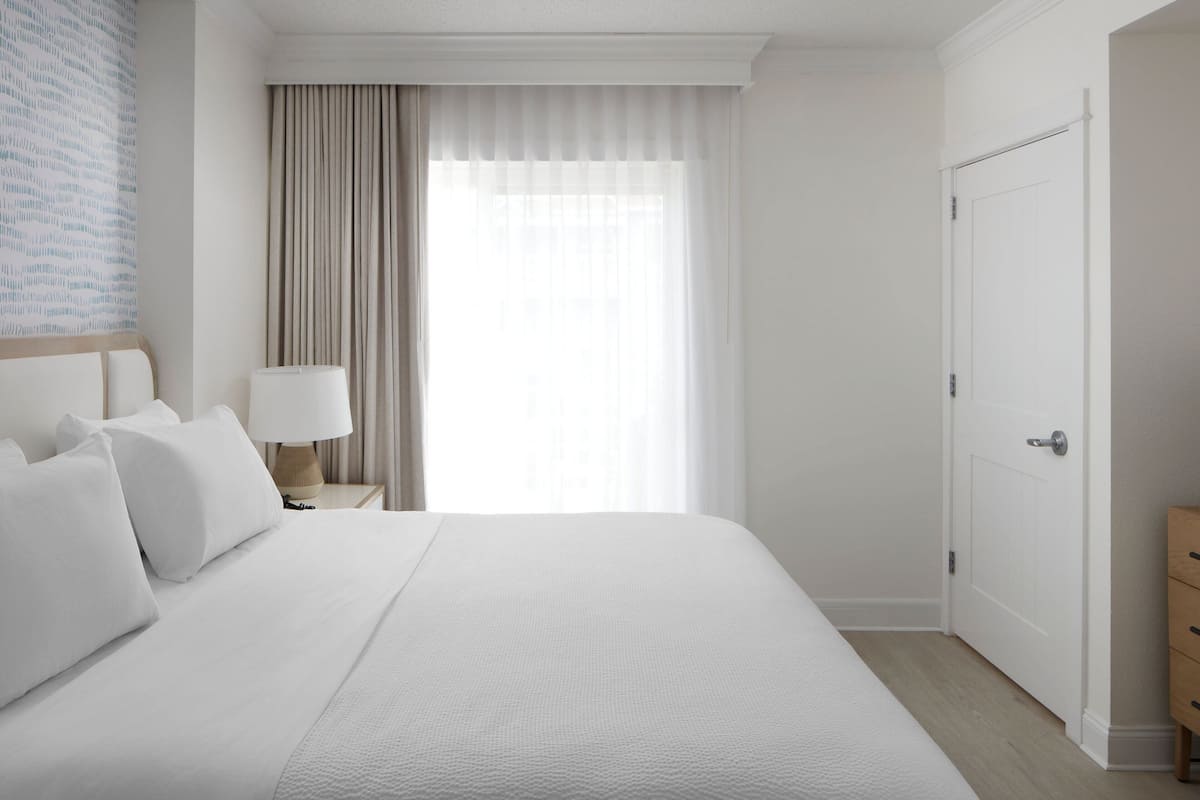 Ein Hotelzimmer mit einem großen Bett, einem Nachttisch mit Lampe, einer Tür und Blick durch ein mit Vorhängen versehenes Fenster hinaus.