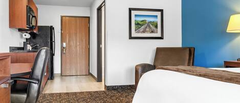 Una habitación de hotel con una cama, un escritorio con una silla, una puerta de madera, un cuadro enmarcado en la pared y una lámpara.