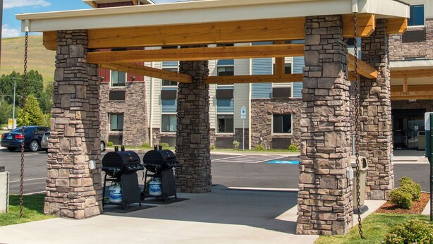 Un espace extérieur abrité, doté de piliers en pierre et d’un toit en bois, avec un barbecue et un parking situé devant un bâtiment avec de nombreuses fenêtres et une enseigne bleue.