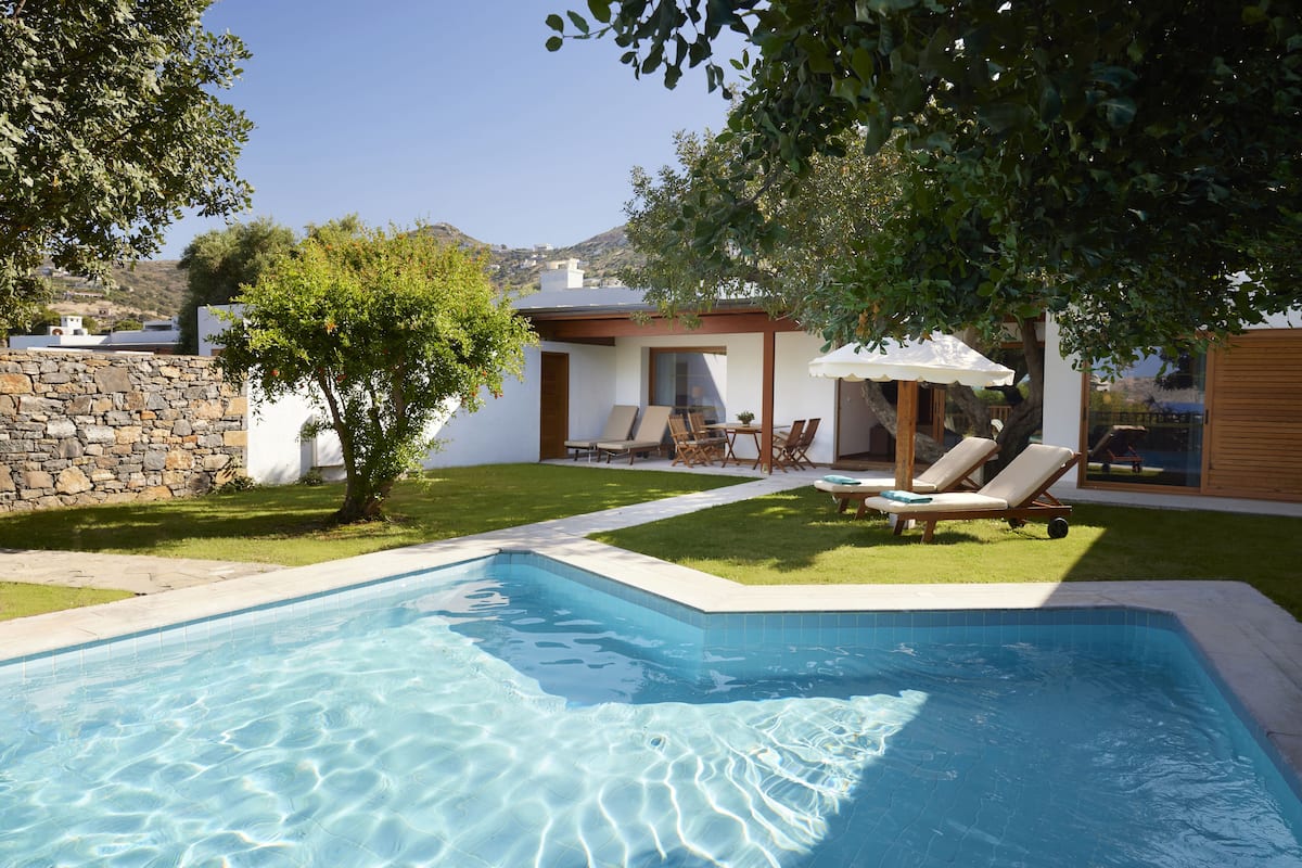 A swimming pool with a stone wall and a small tree in the foreground. There is a patio area with a table and chairs, and a building with a white exterior in the background.
