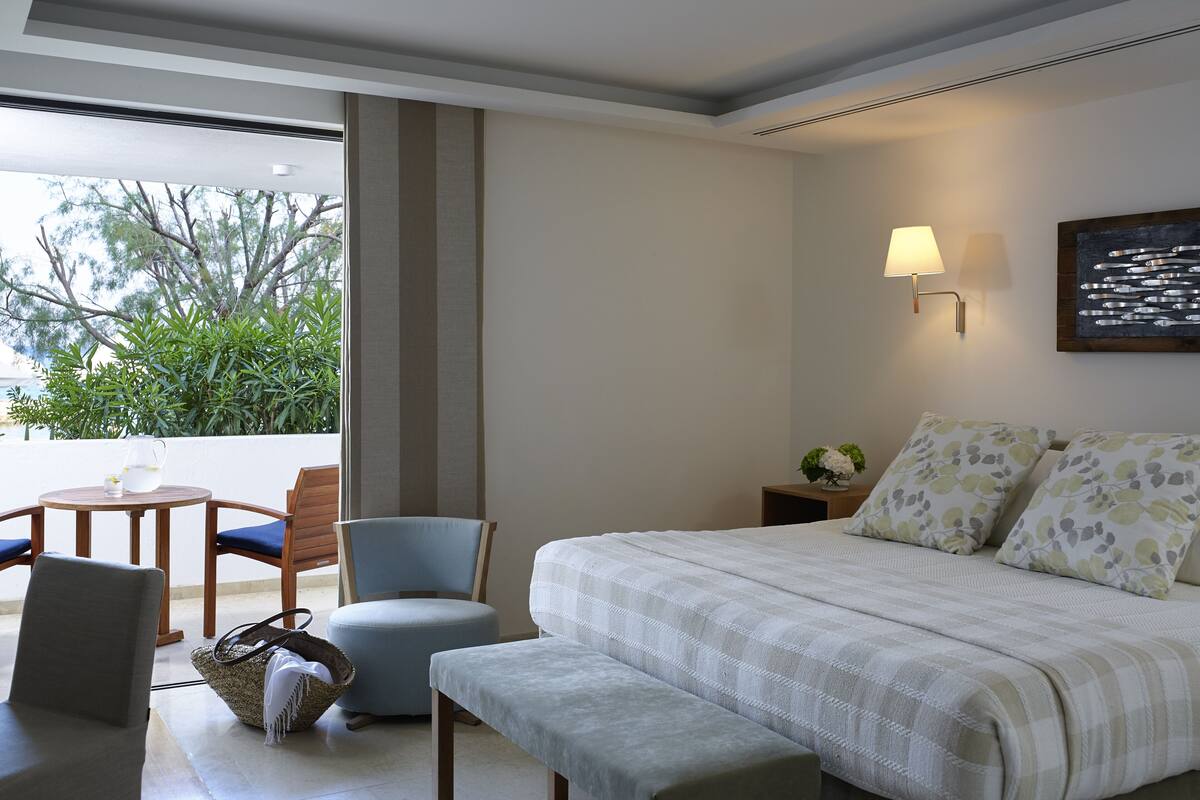 A modern hotel room with a large bed, a seating area with a table and chairs, and a view of greenery outside.