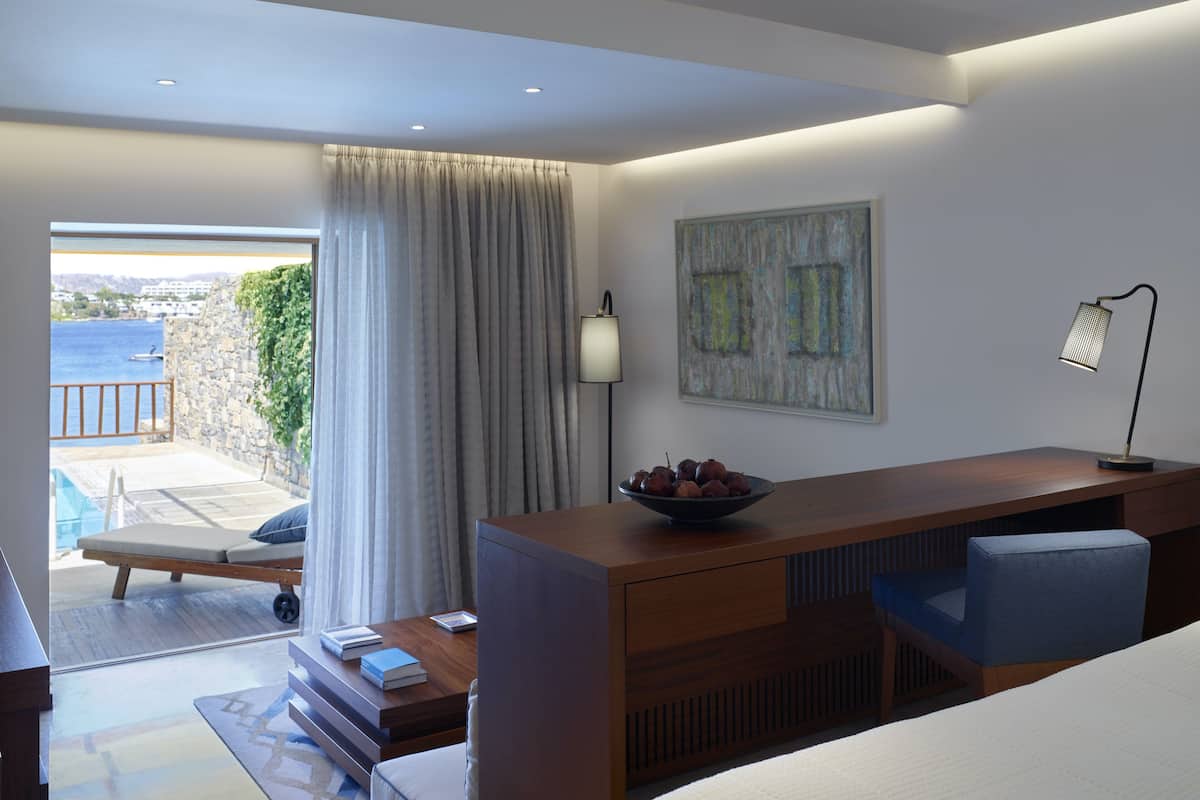 A modern hotel room with a bed, a desk with a bowl of fruit, a lamp, and a view of the sea.