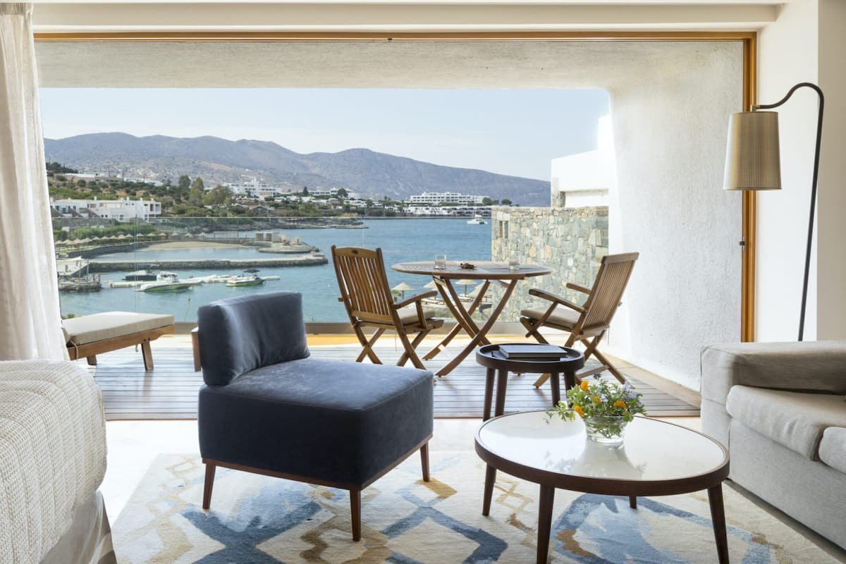 A balcony with a view of a harbor, a round table, two chairs, a sofa, and a lamp.