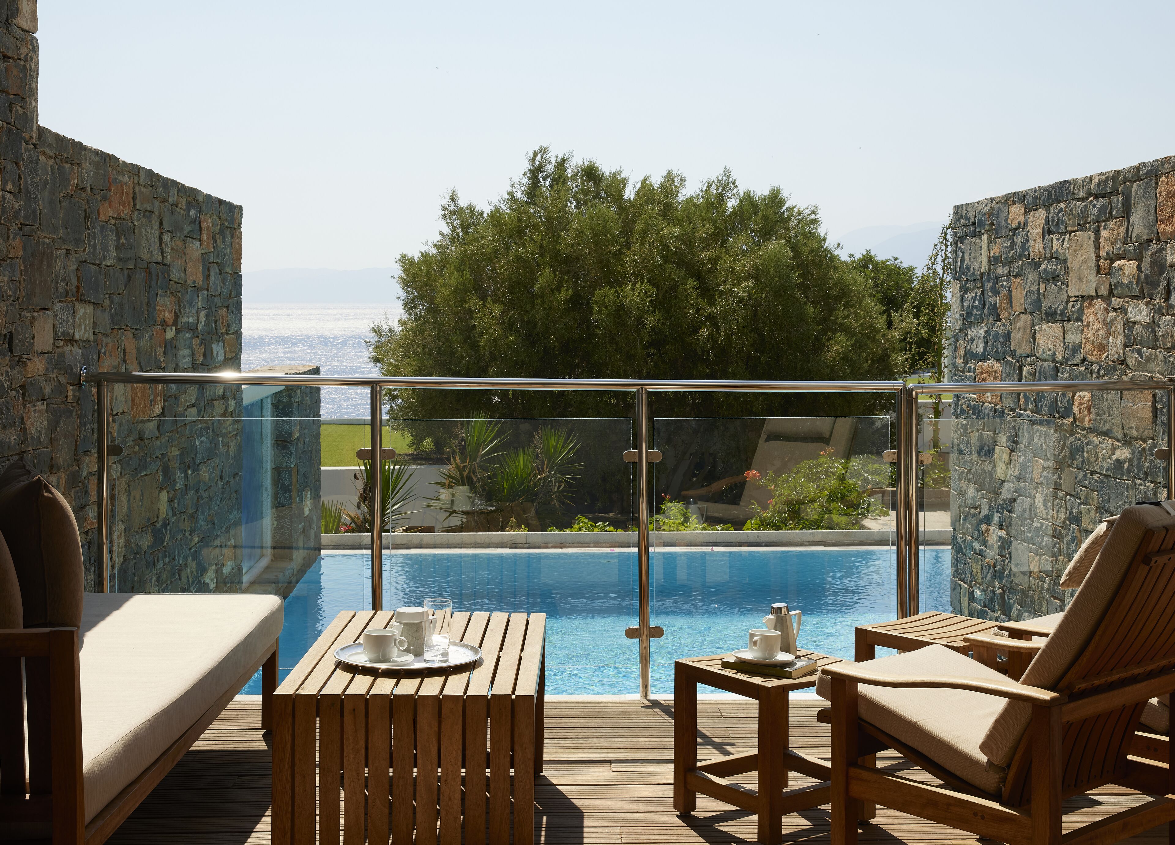 Eine Holzterrasse mit Pool, Liegestühlen und einem Glasgeländer, das einen Blick auf eine Landschaft mit Bäumen und eine Ferne bietet.