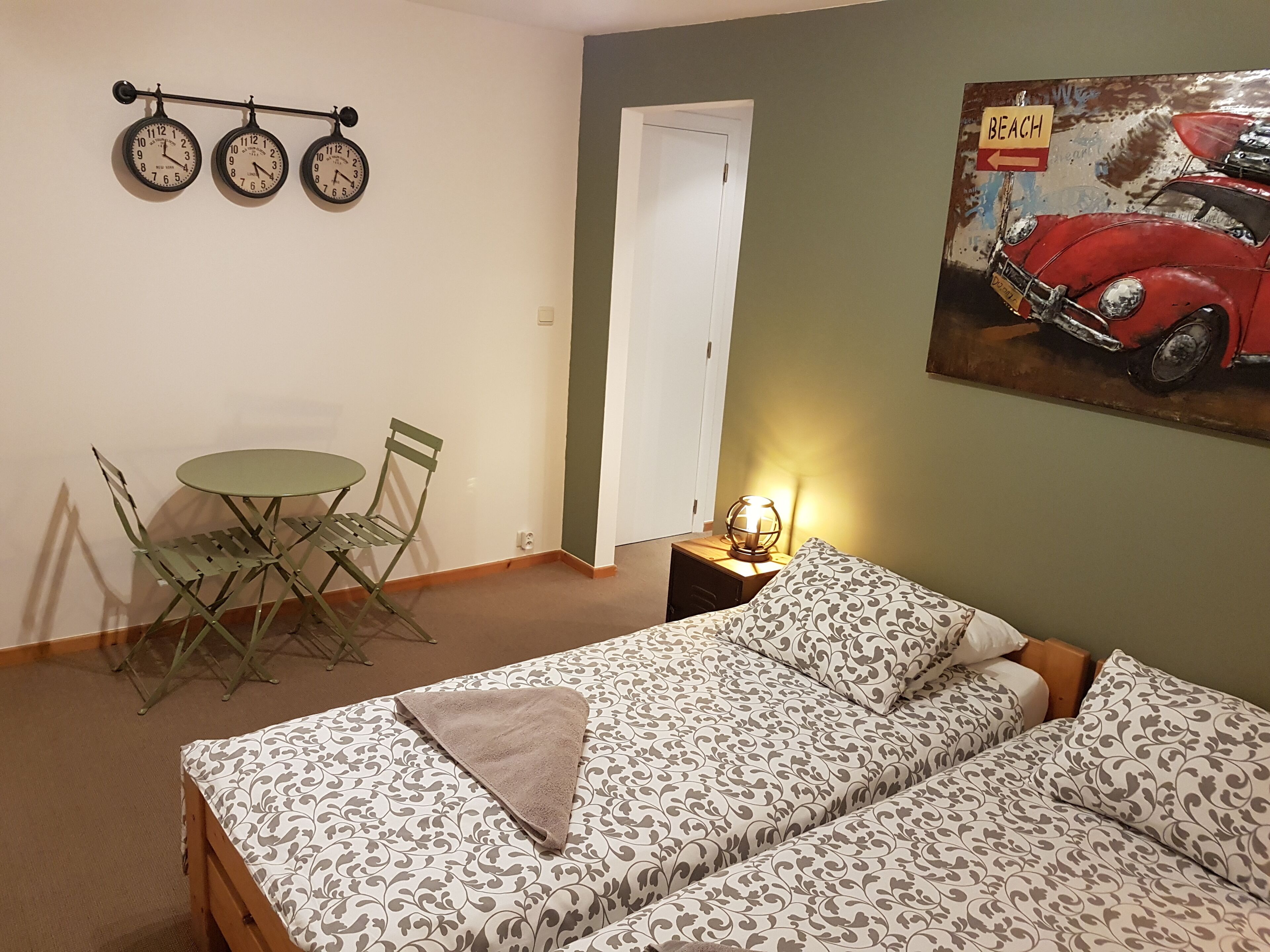 Een kamer met een bed, een klein tafeltje met stoelen, een wandklok en een schilderij van een auto.