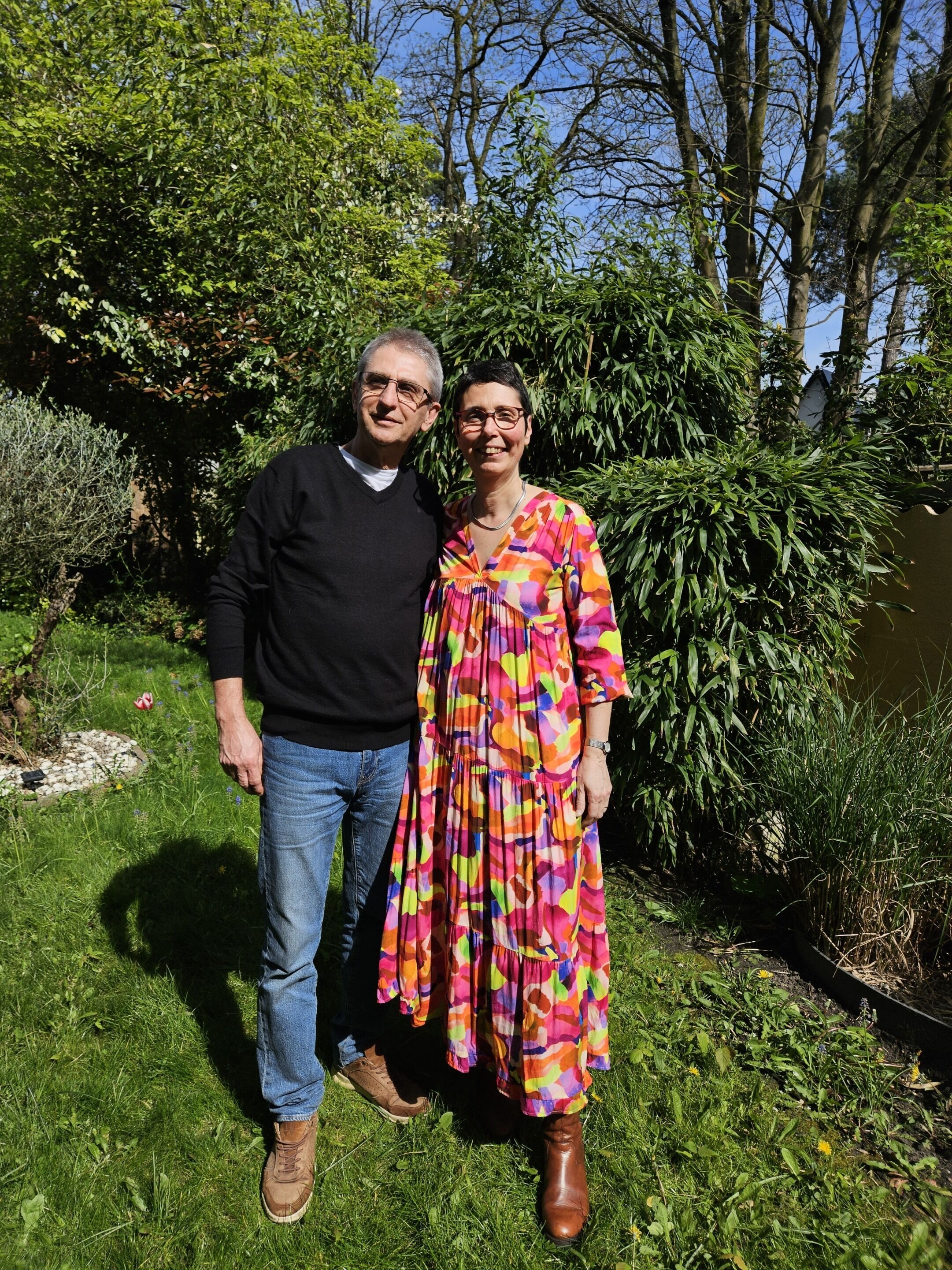 Twee personen staan in een tuin met weelderige begroeiing en bomen op de achtergrond.