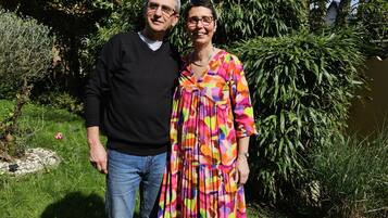 Twee personen staan in een tuin met weelderige begroeiing en bomen op de achtergrond.