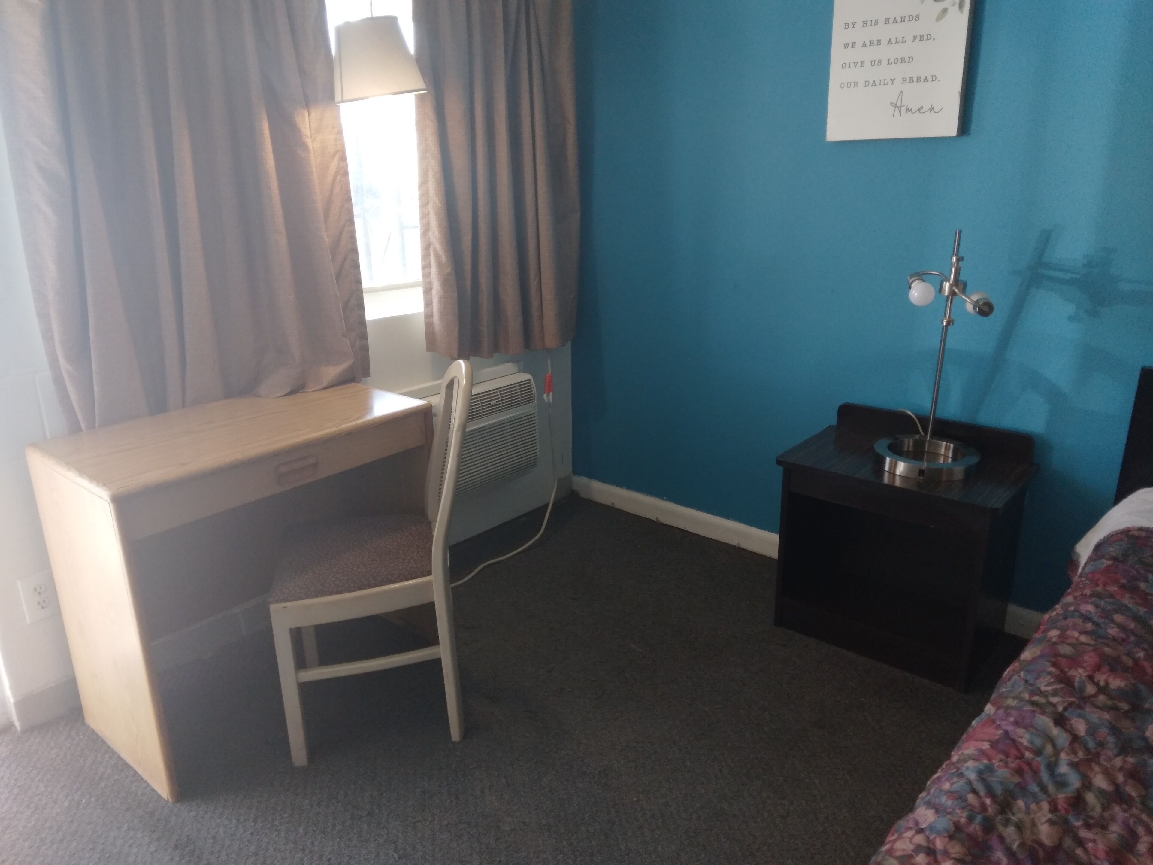 Habitación de hotel con escritorio, silla, cama, mesita de noche, lámpara y una pared con un texto religioso.