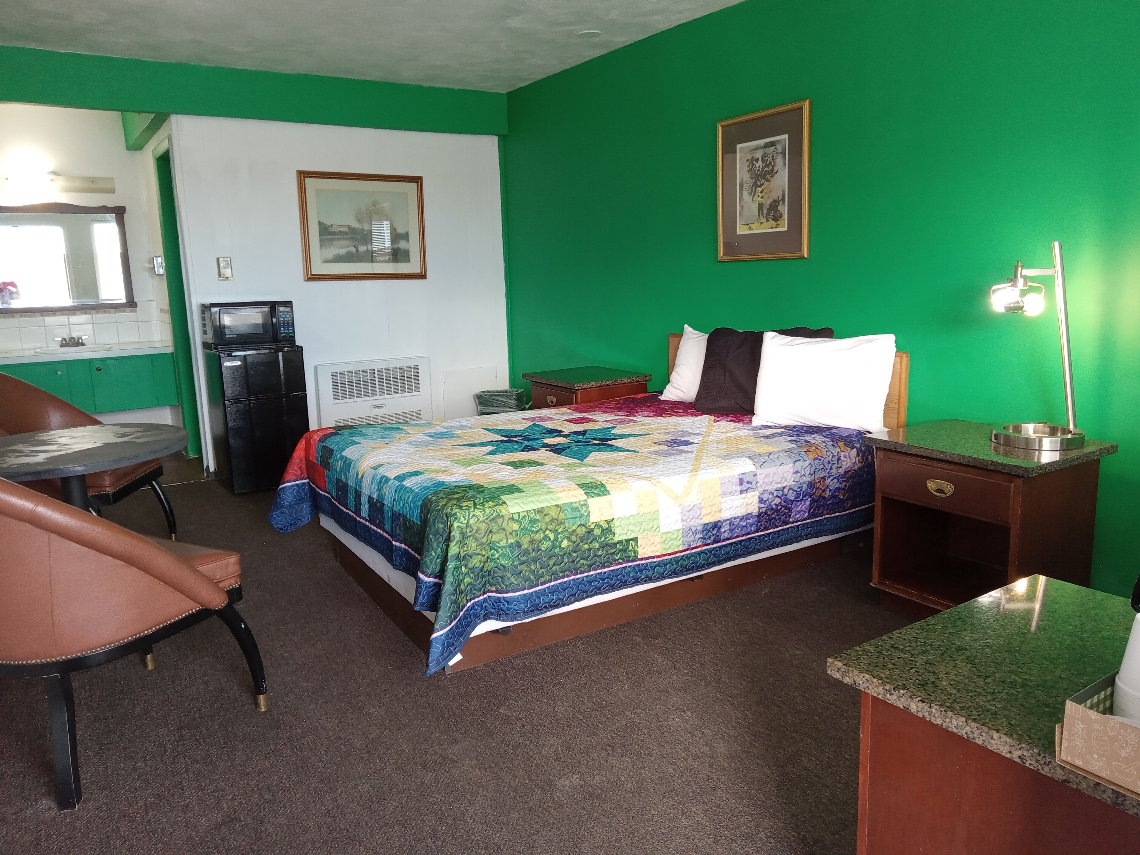 A hotel room with a bed, a desk, a chair, a small kitchenette, and a green wall.