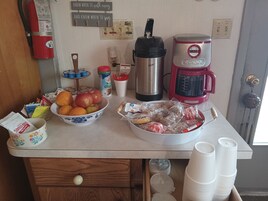 Una encimera de cocina con una cafetera, un termo, un cuenco de fruta y diversos utensilios de cocina.