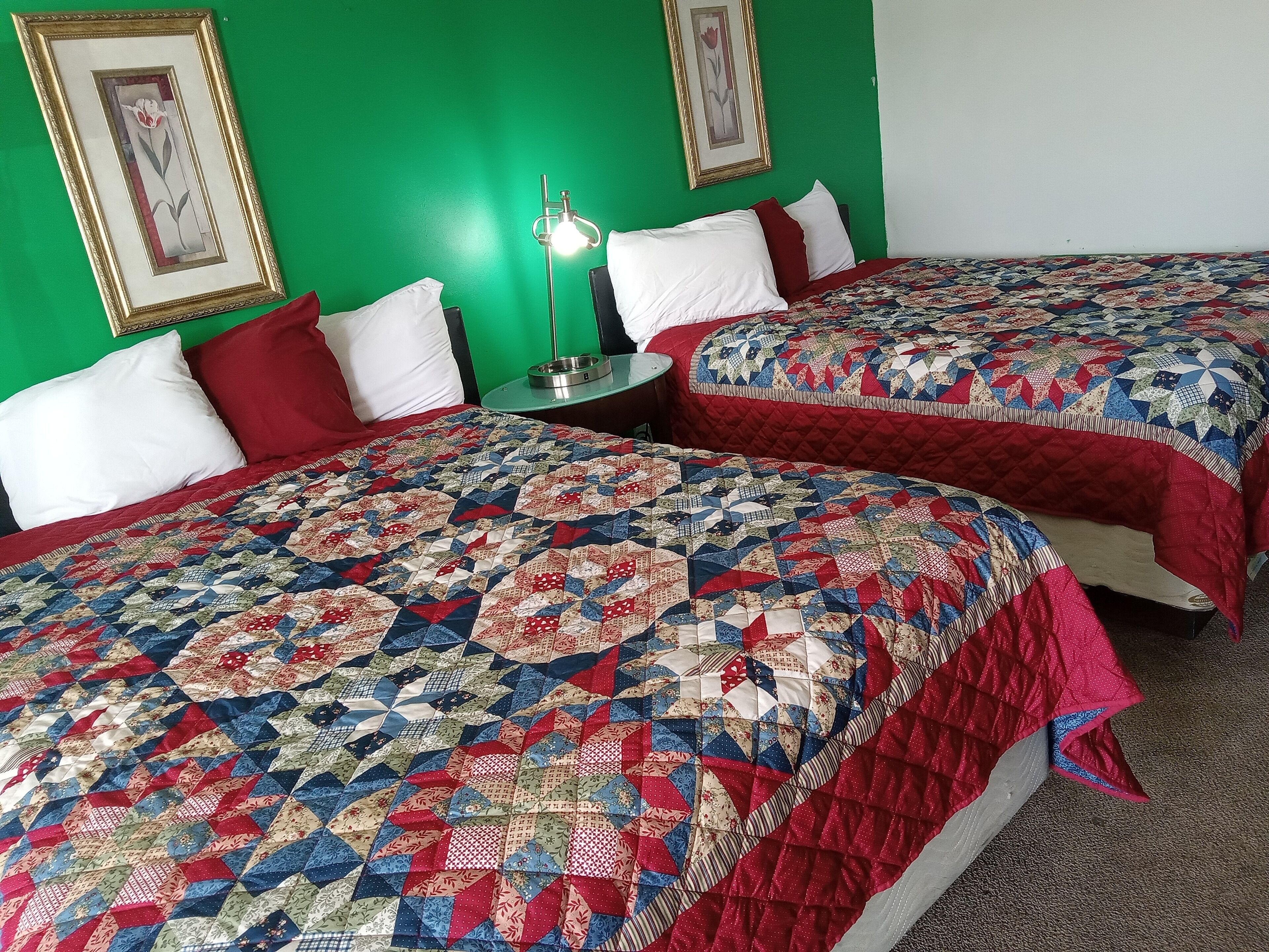 Habitació d'hotel amb dos llits, cada un amb una colcha vermella i blanca. Hi ha quadres emmarcats a les parets verdes, una taula petita amb una làmpada i un almoiner blanc en un dels llits.